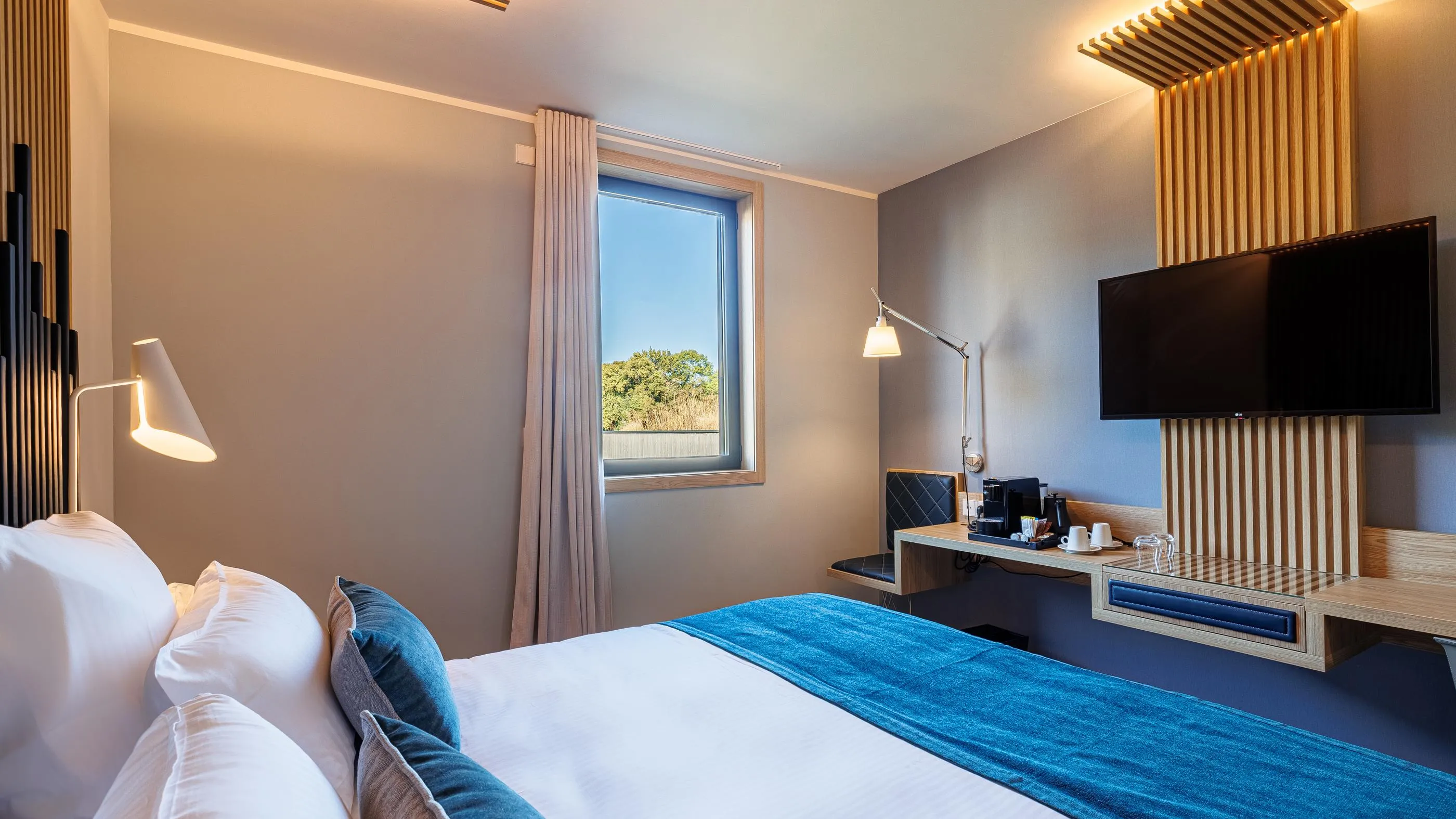 Hotel room with a double bed, a window, a desk with a coffee machine, and a flat-screen TV mounted on the wall.