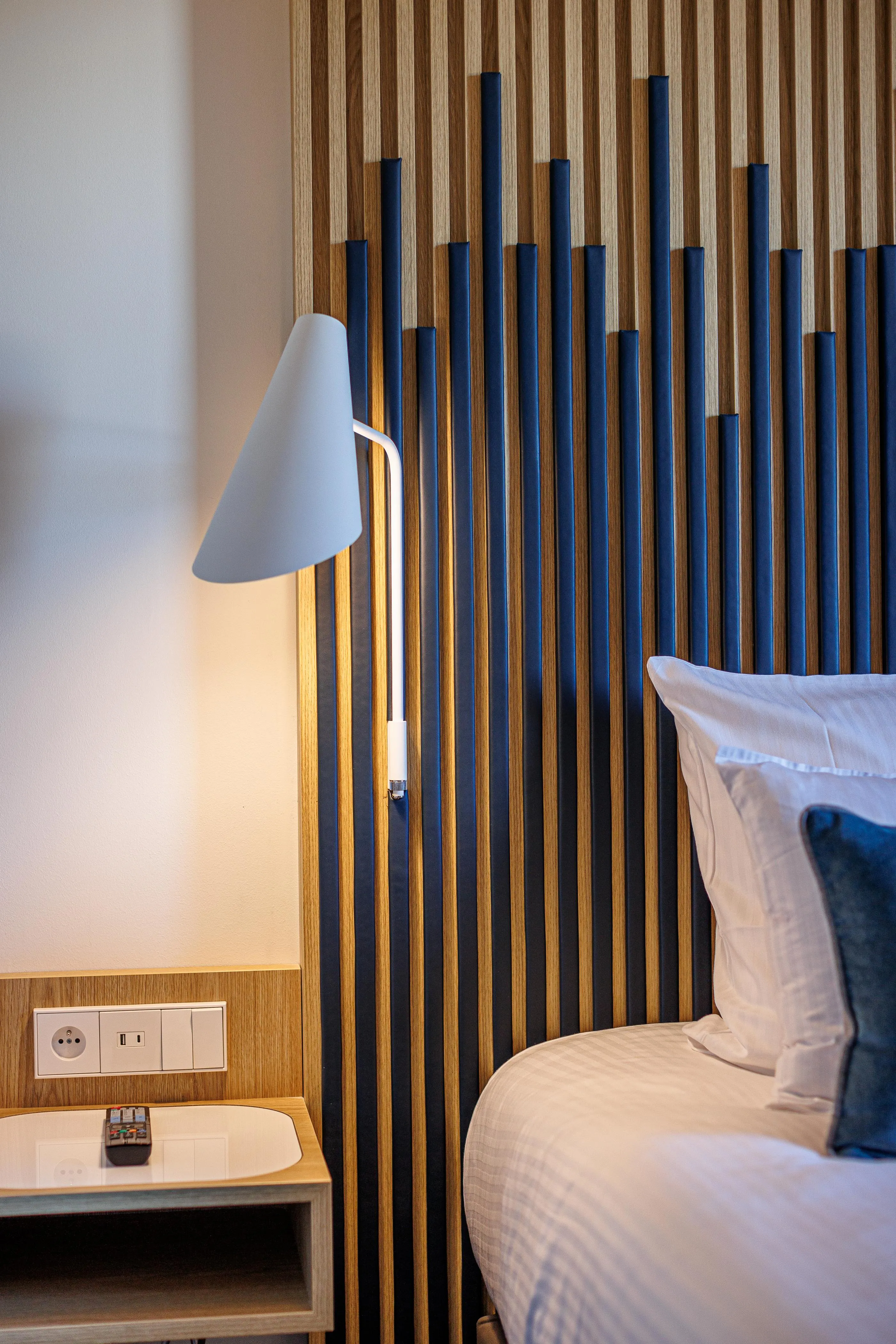 Bed with a wooden and metal headboard, a lit lamp turned on, a nightstand with an electrical outlet and a remote control