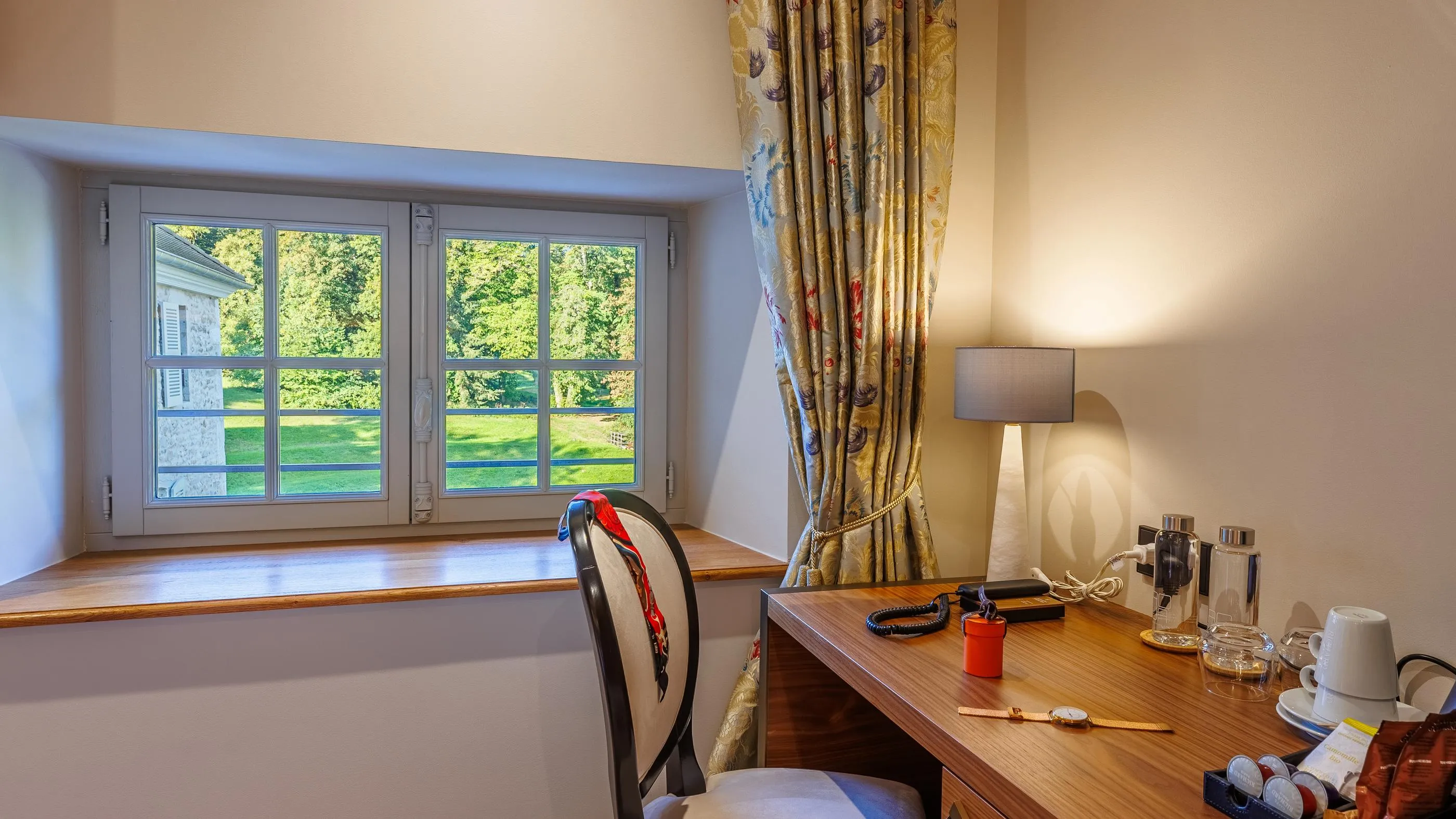 Wooden desk with a lit lamp, a cup, glasses, and a window overlooking a tree-lined garden.