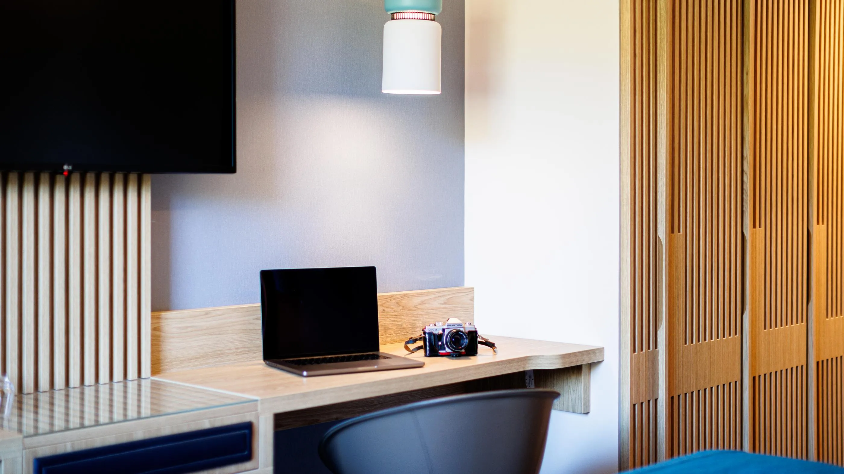 A wooden desk with a laptop and a camera, under a TV mounted on a vertically slatted wall