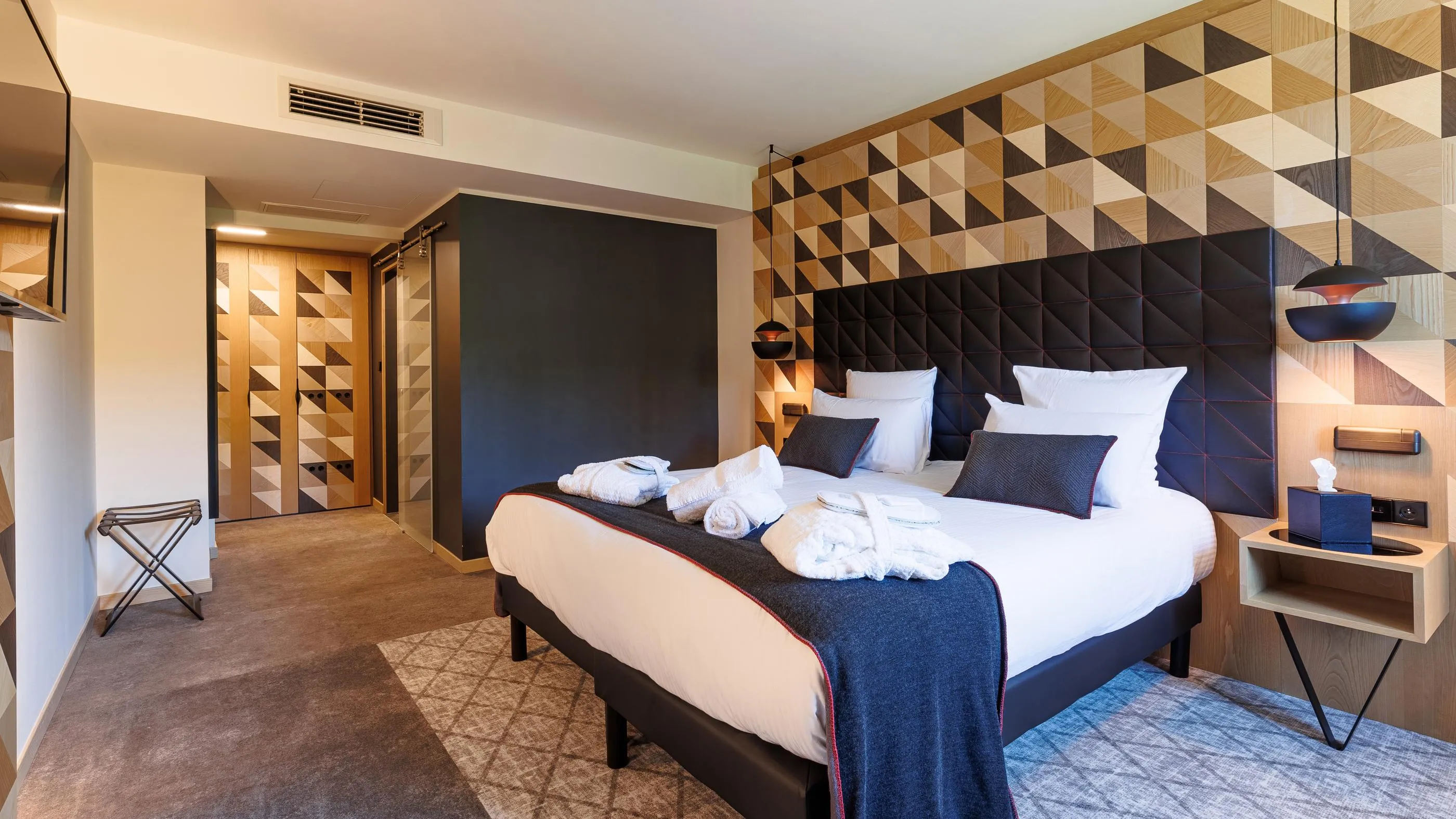 Modernes Hotelzimmer mit Doppelbett, geometrischem Holzkopfteil, Designleuchten und offener Tür zu einem ähnlichen Flur