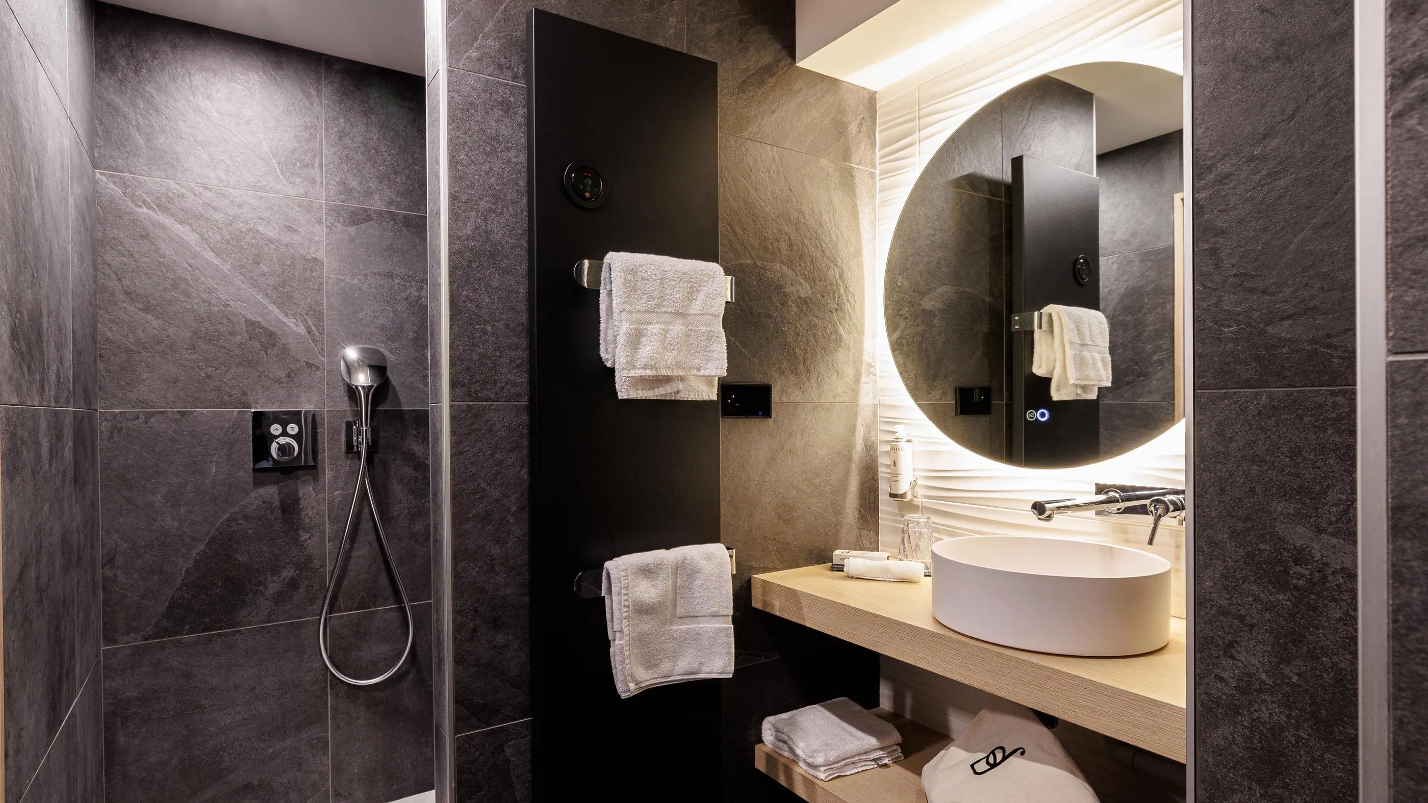 Moderne Badezimmer mit Handbrause, rundem Spiegel, weißem Waschbecken und Handtüchern