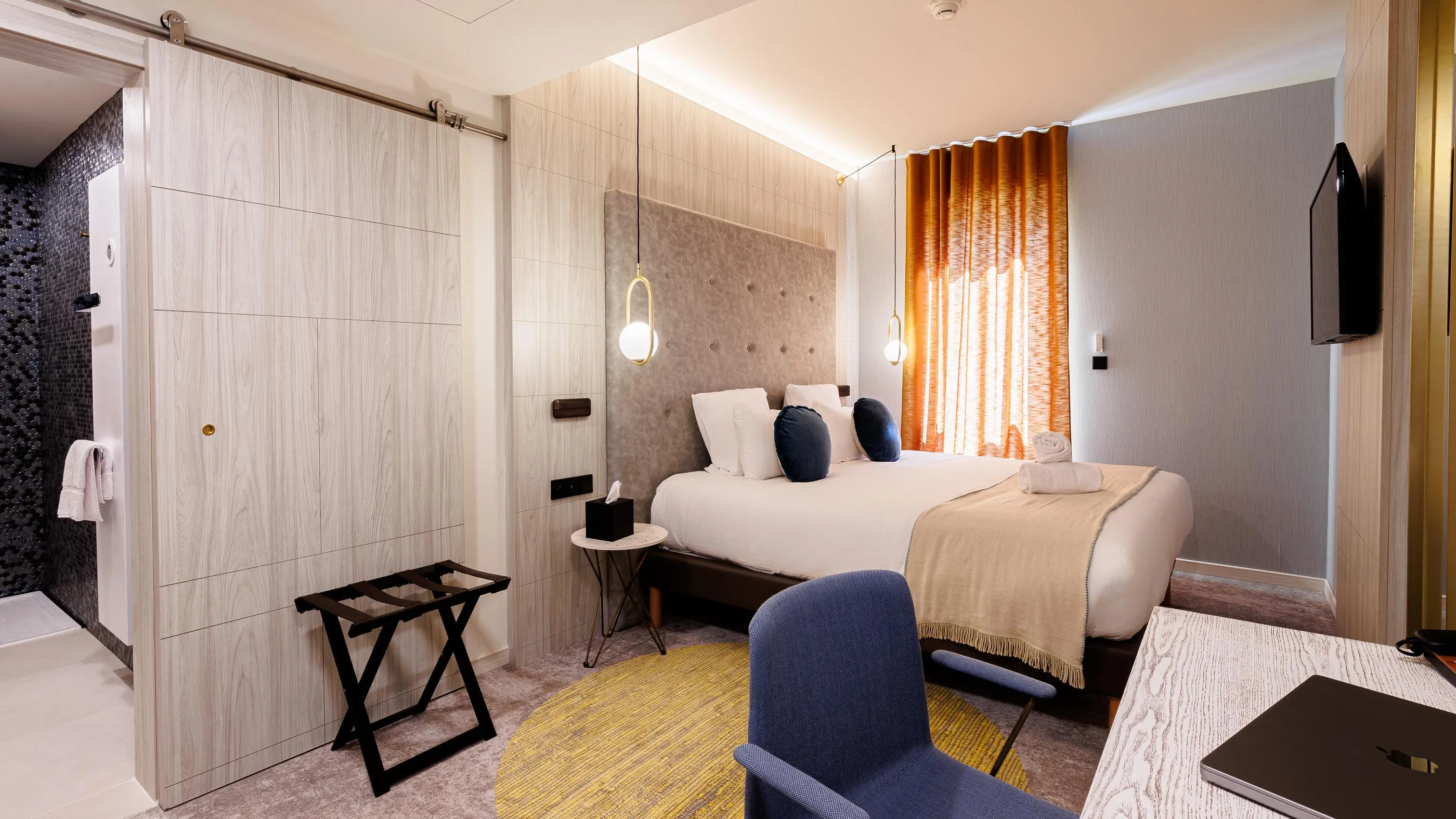 Hotelzimmer mit einem Bett, einem Nachttisch, einem Sessel, hängenden Lampen und einer offenen Badezimmertür