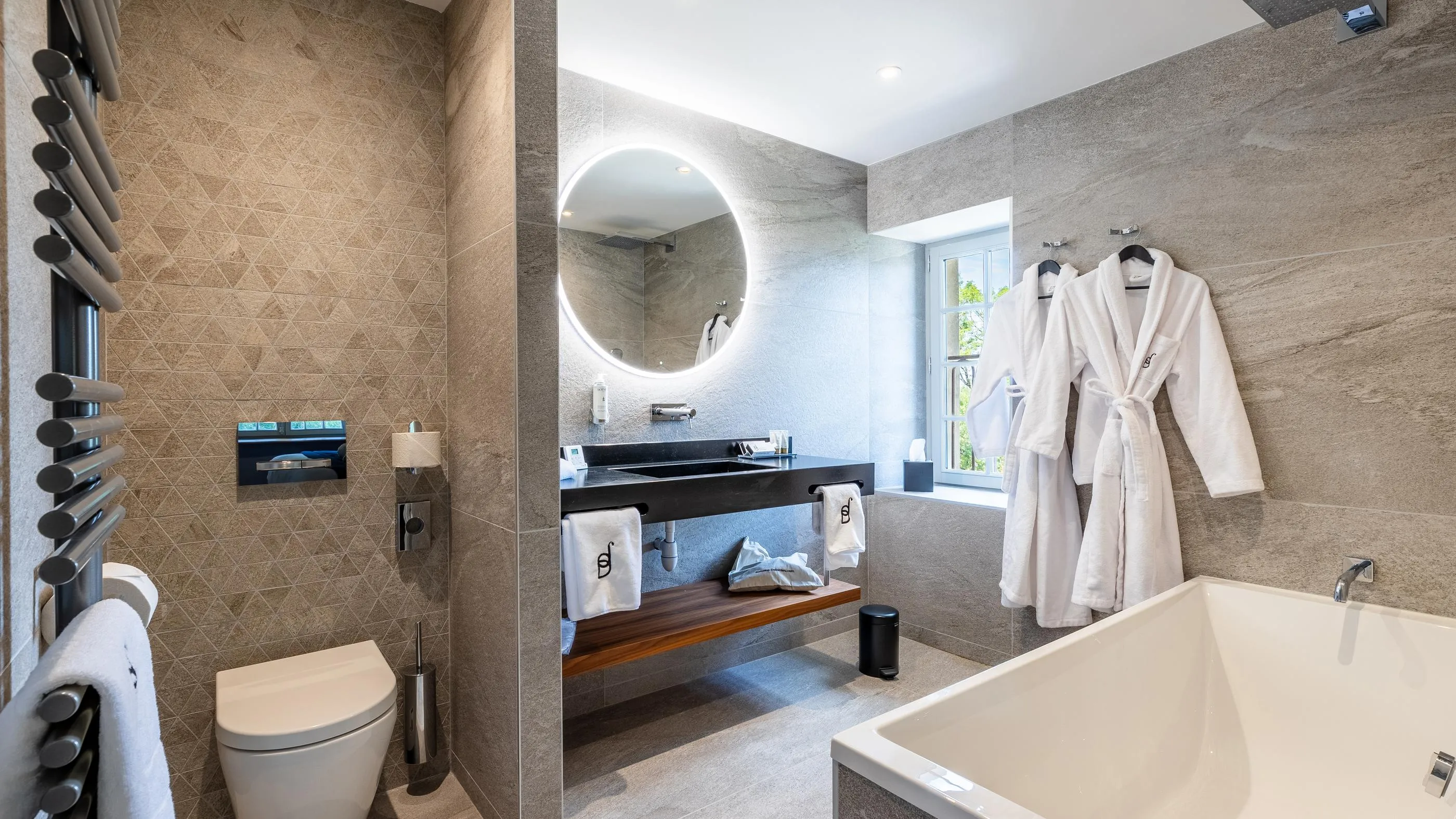 Moderne Badezimmer mit Badewanne, Toilette, Waschbecken, rundem Spiegel und weißem Bademantel