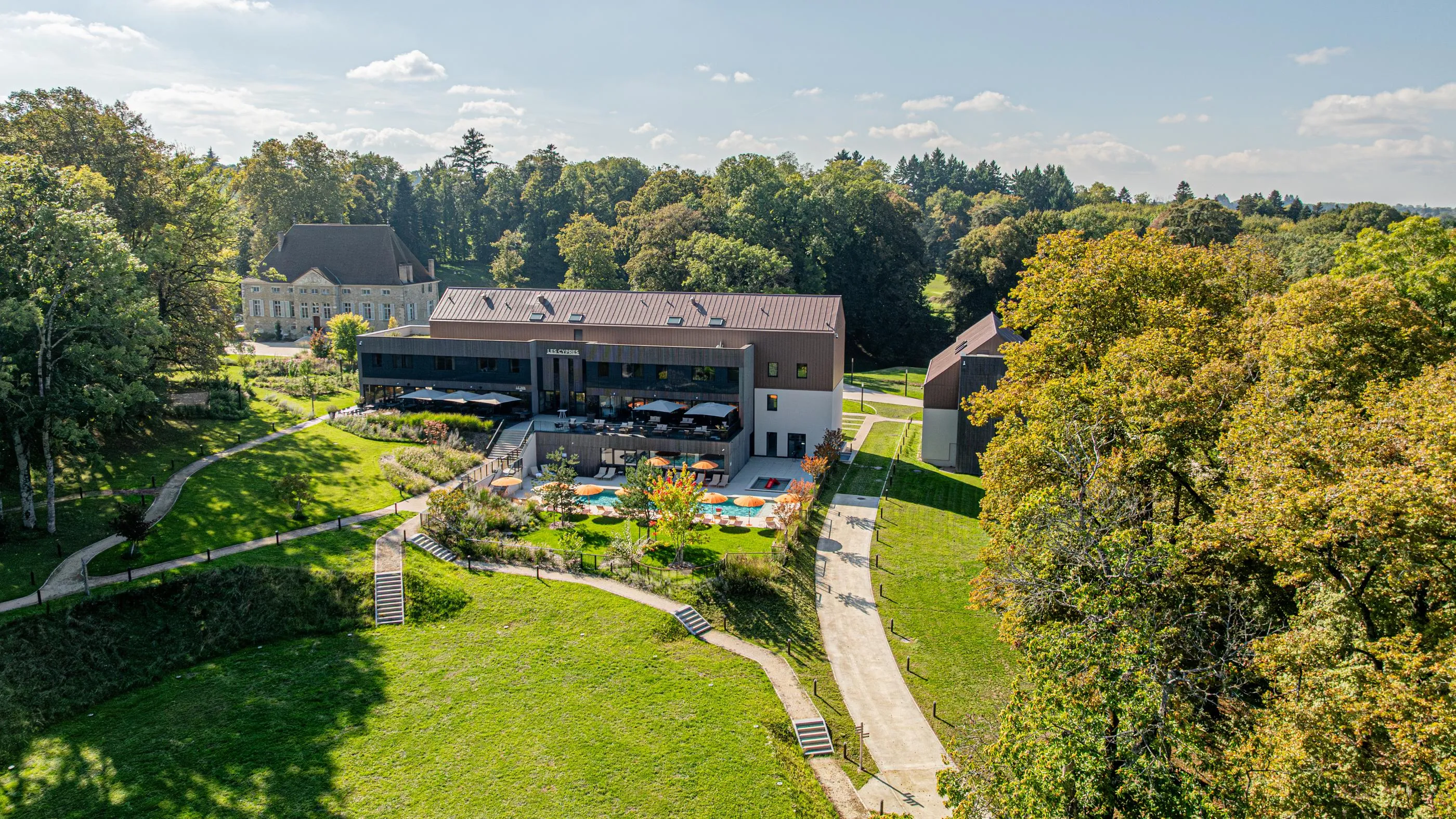 Un bâtiment moderne entouré d'arbres et de pelouses, avec une terrasse et une piscine, situé près d'un parc arboré et d'un autre bâtiment plus ancien