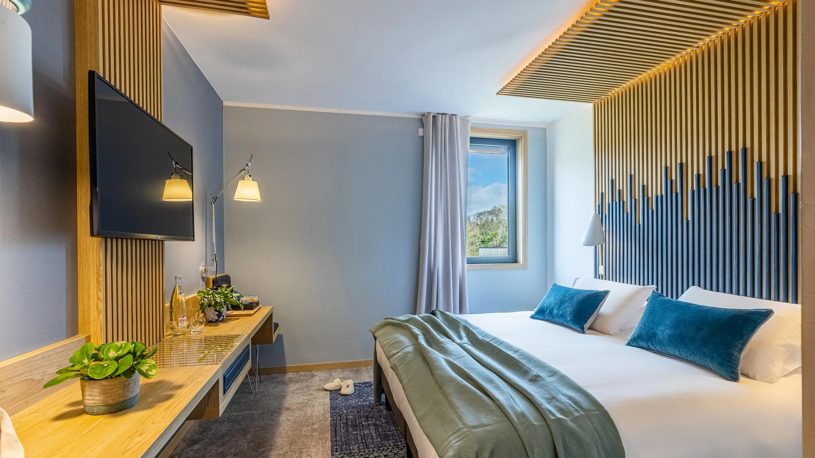 Modern hotel room with a double bed, a flat-screen TV, a desk, and a window letting in natural light.