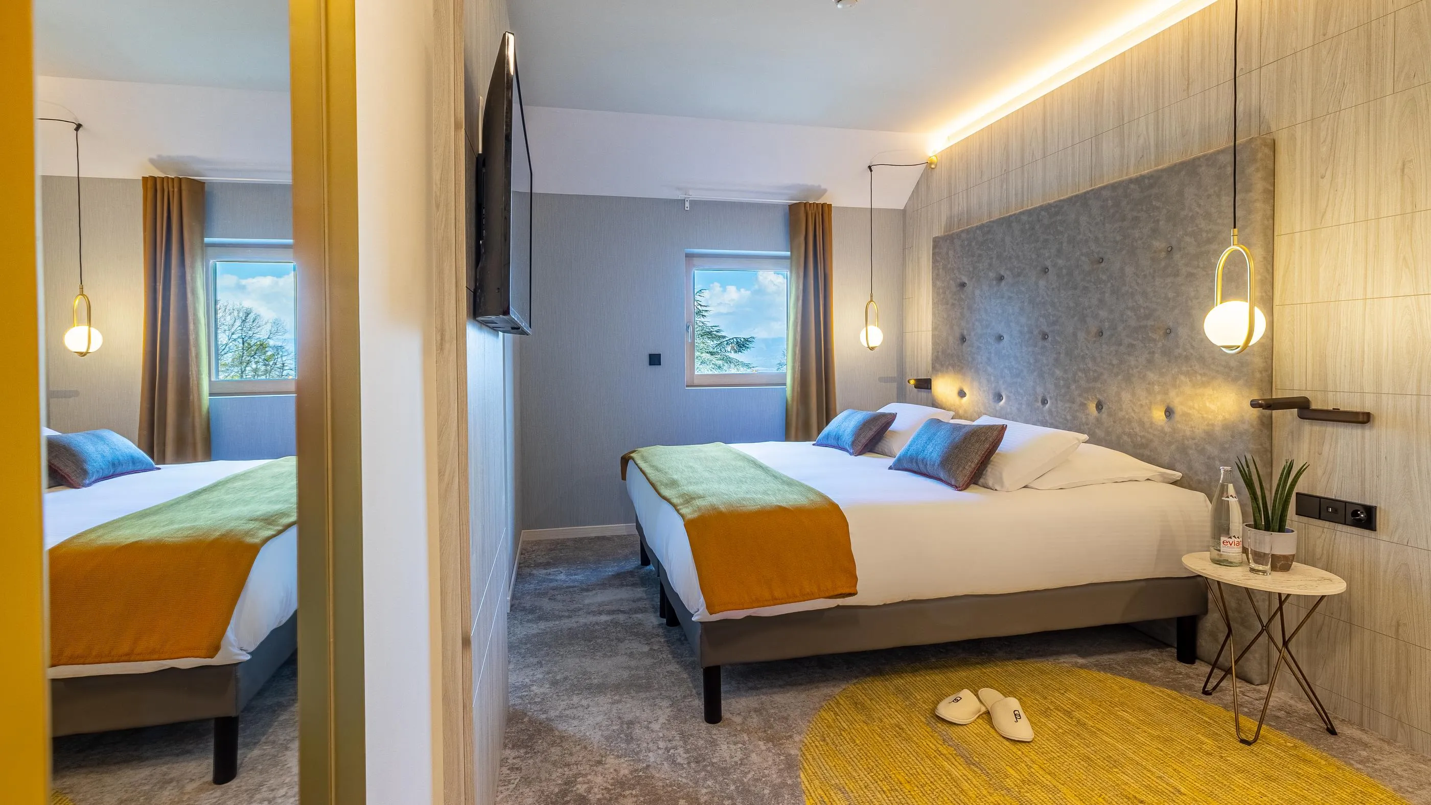 Hotelzimmer mit zwei Einzelbetten, einem Nachttisch, hängenden Lampen und einem Blick nach außen durch die Fenster