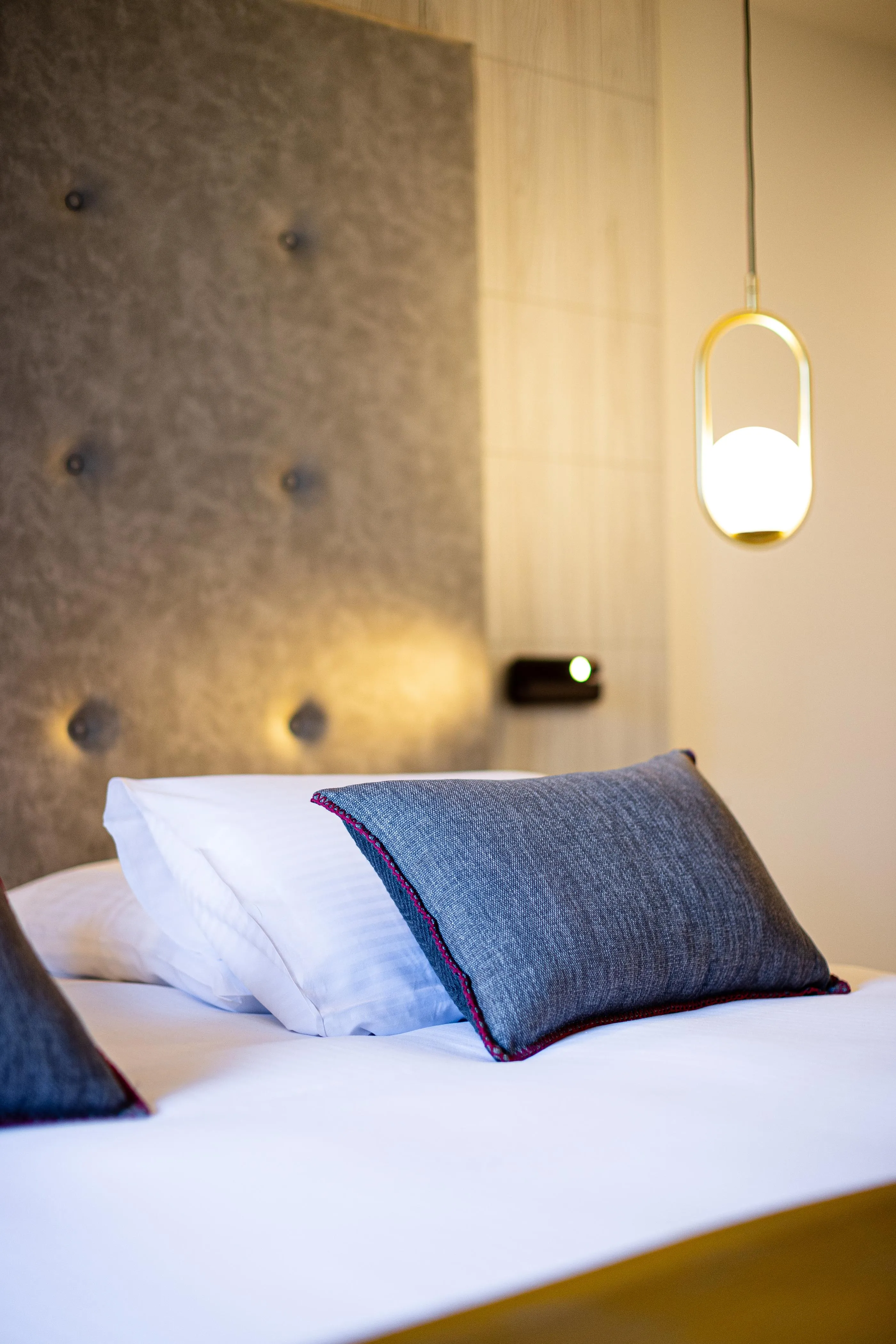Bett mit zwei Kissen, davon eines grau mit roter Umrandung, und einer Lampe, die rechts hängt