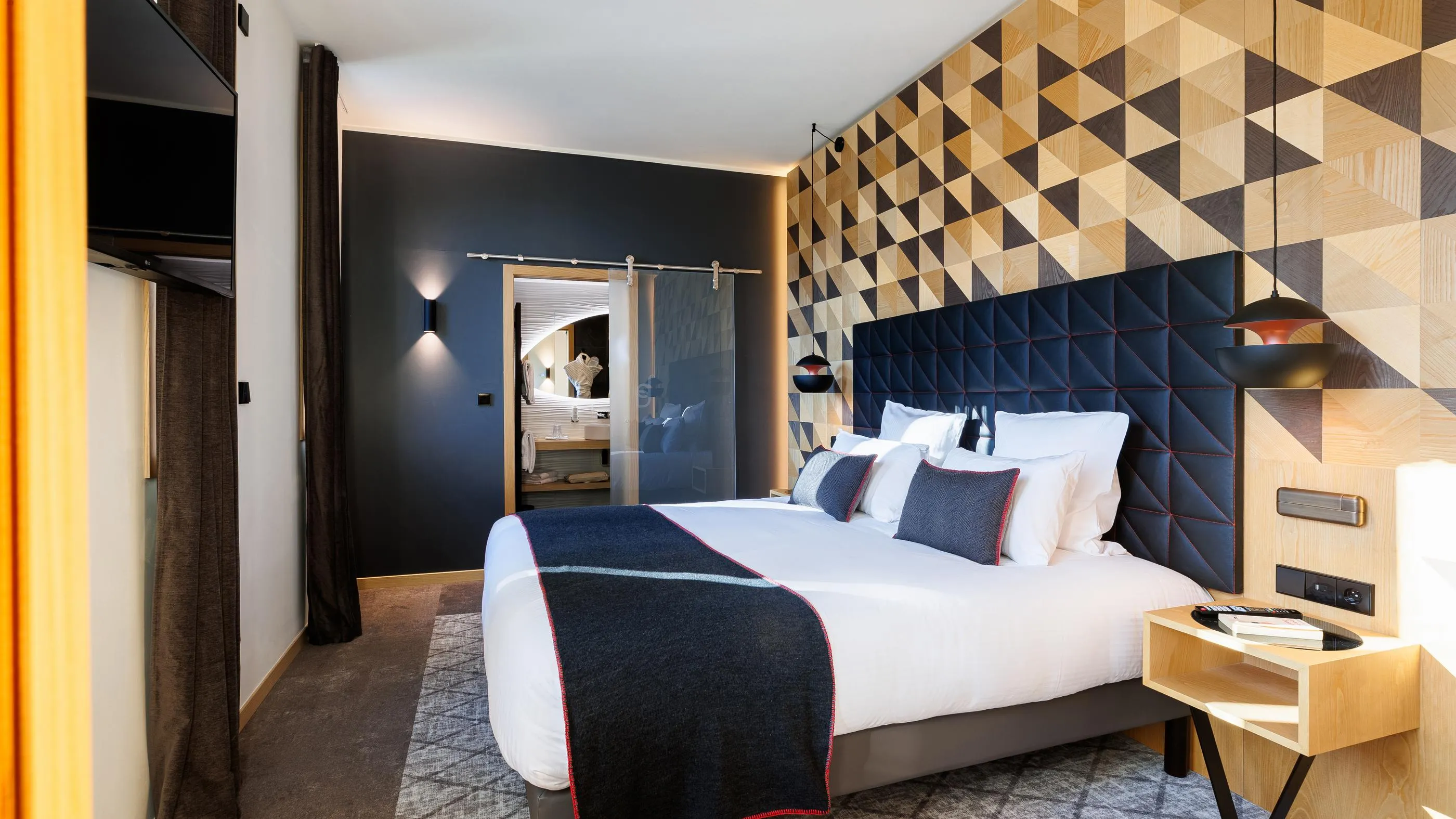 Modernes Zimmer mit einem Doppelbett, einem dunkelblauen Samtkopfteil und einer mit hellen und dunklen geometrischen Holzmustern verzierten Wand