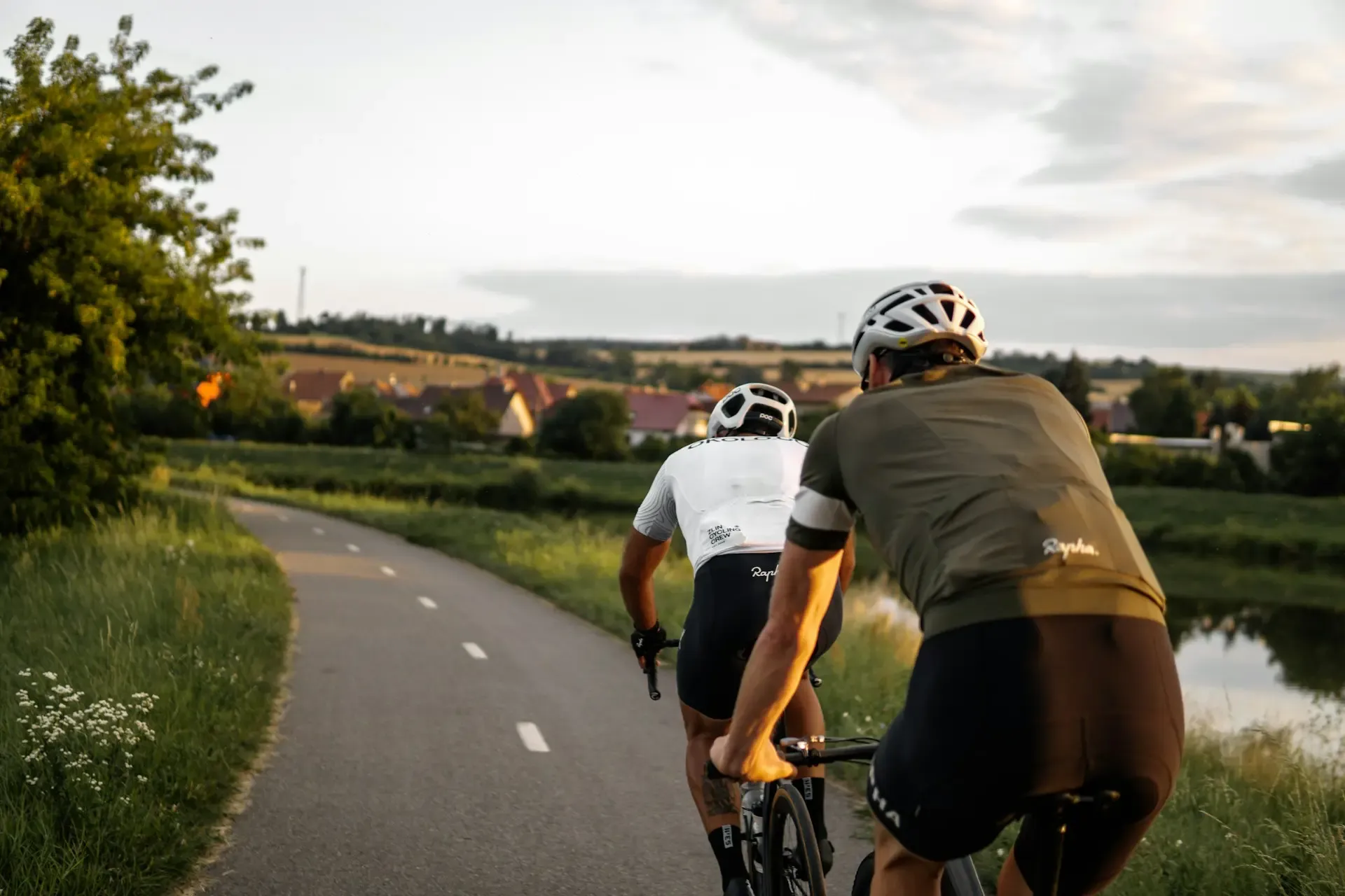 Deux cyclistes portent des casques et des tenues de sport, roulant sur une route bordée d'herbe et d'arbres, avec un village et des champs en arrière-plan