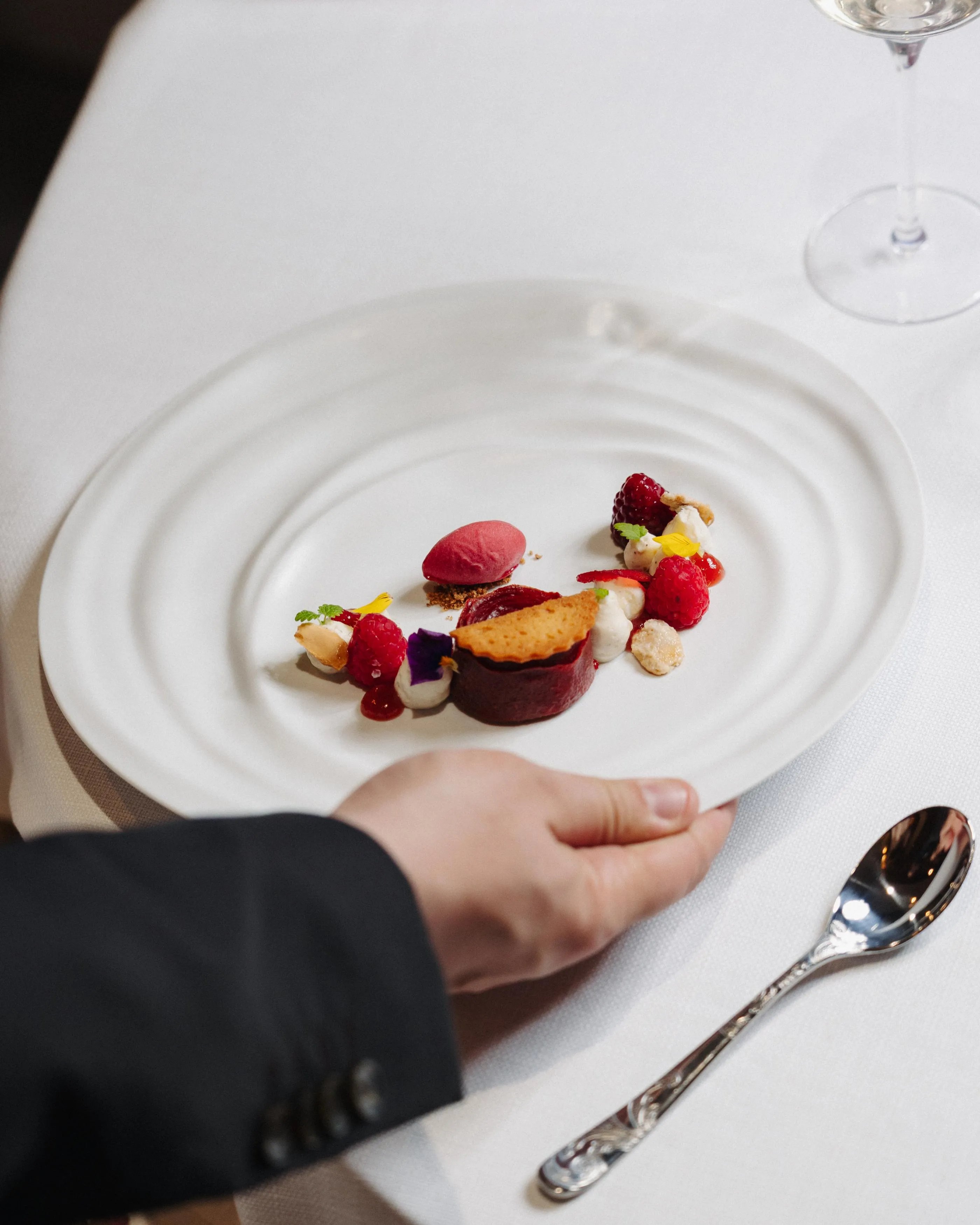 Une main tenant une fourchette s'approche d'une assiette contenant un plat composé de divers éléments colorés, posé sur une nappe blanche avec une cuillère à côté