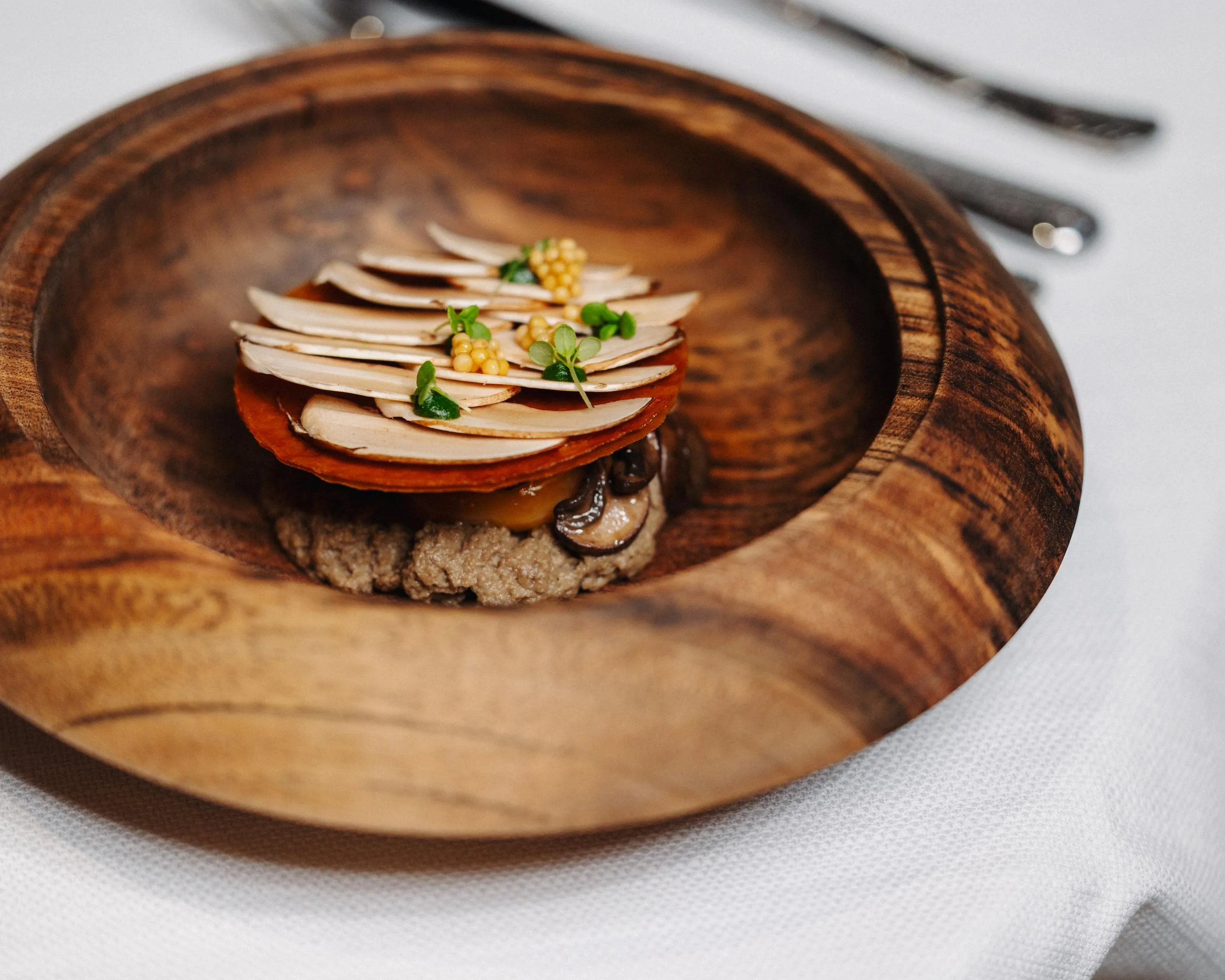 A wooden dish containing a stack of thin vegetable slices, mushrooms, and seeds, placed on a white tablecloth.