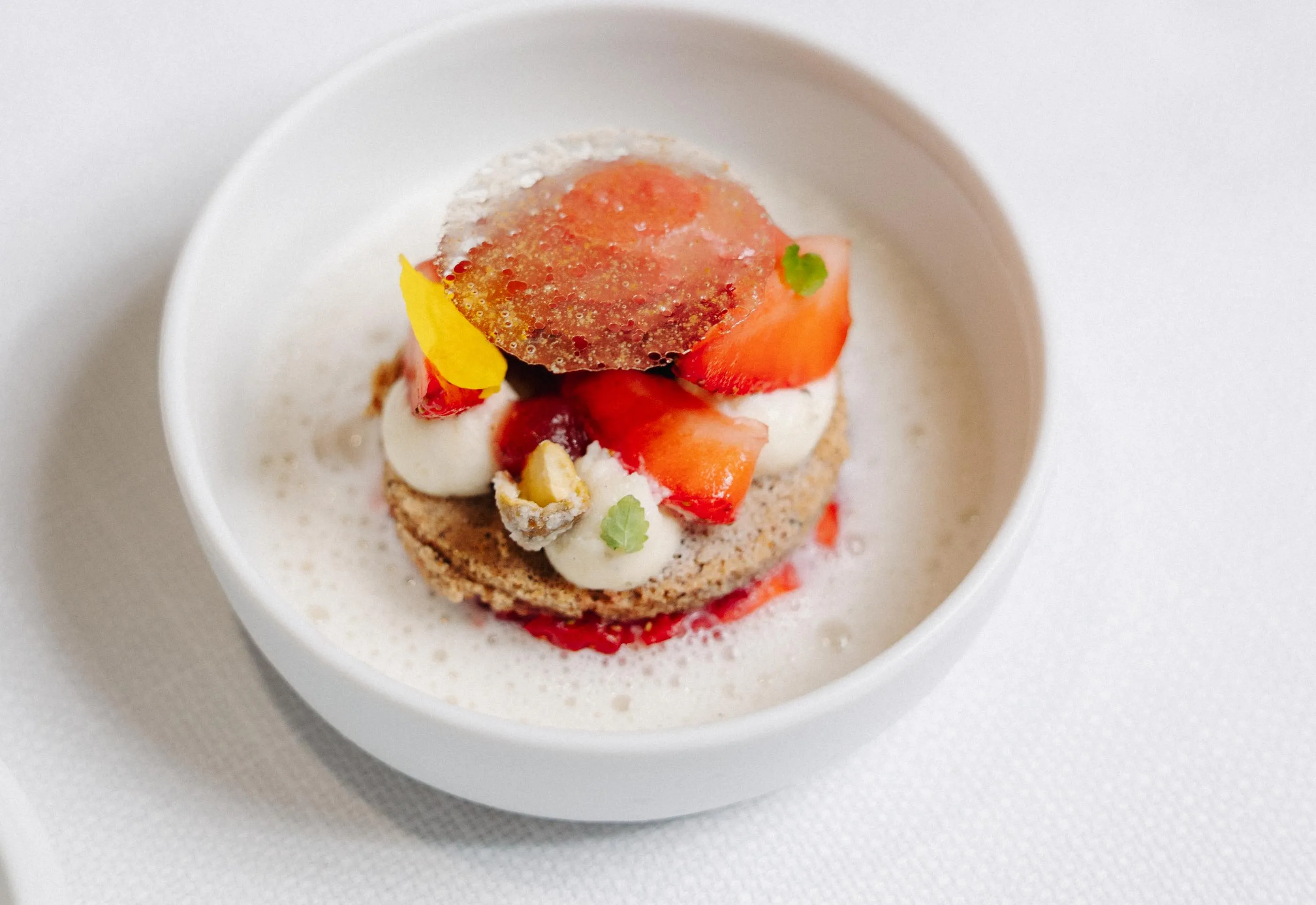 Un dessert dans un bol blanc composé d'une base de biscuit, de mousse, de morceaux de fraise et d'une fine tranche de fraise caramélisée