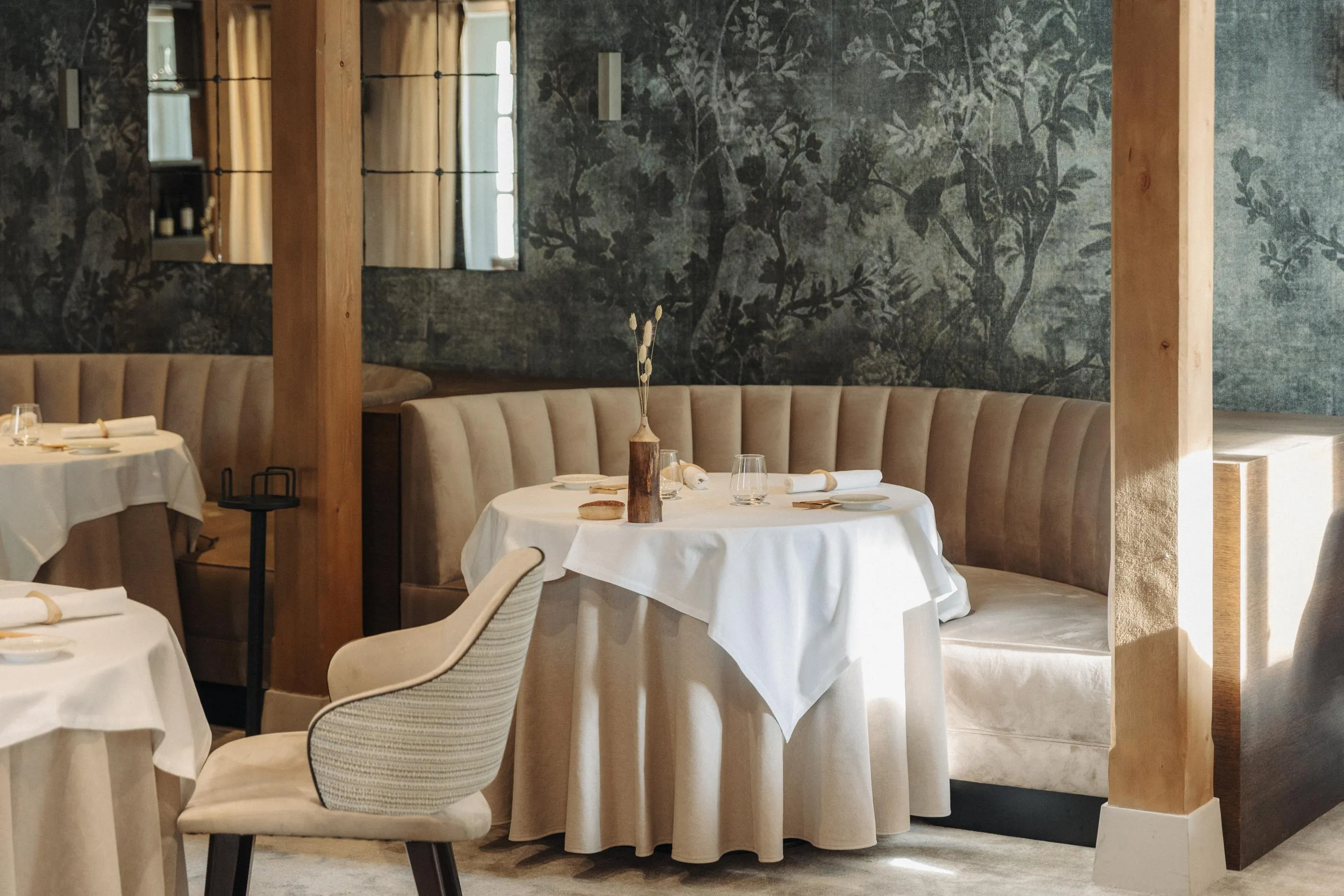 A table set with glasses and floral decorations in a restaurant with walls adorned with plant motifs and padded benches.