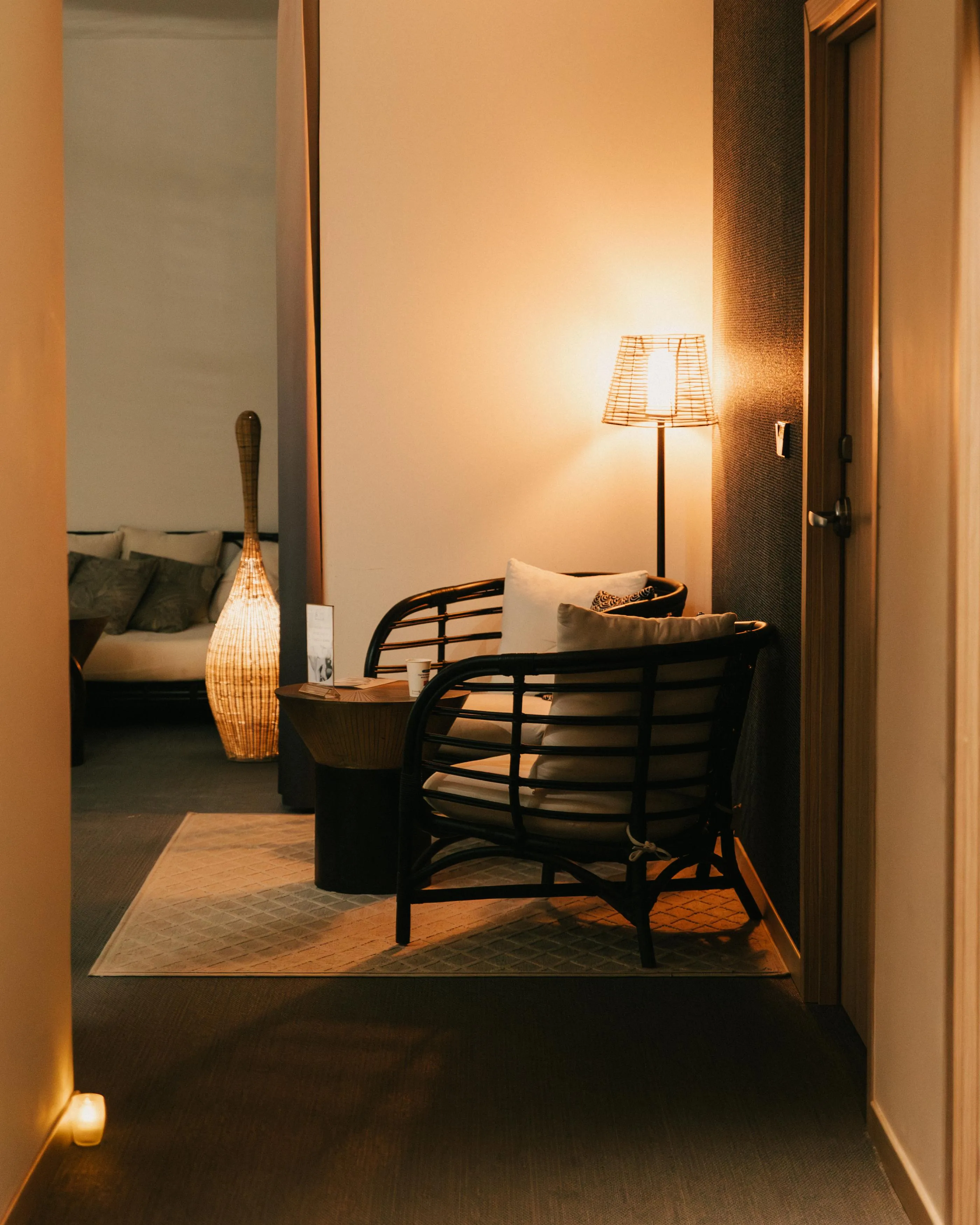 Un fauteuil avec des coussins et une lampe allumée dans une pièce faiblement éclairée, vue à travers une porte ouverte