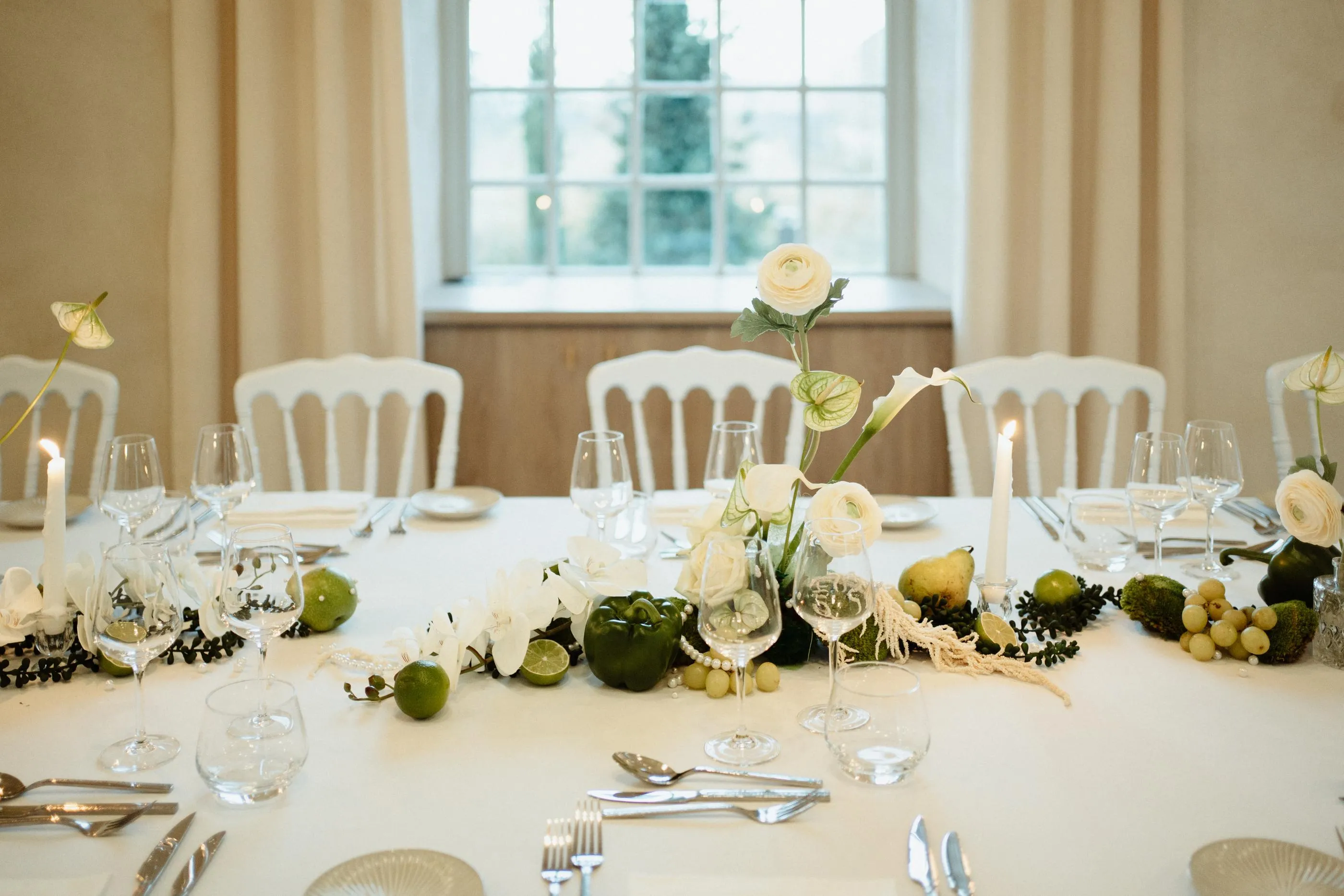 Table dressée avec des verres, des couverts, des bougies et des fleurs blanches et vertes au centre, entourée de chaises blanches